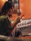 Вид збоку на стильну вдумливу жінку, яка серфінгує мобільний телефон і п'є каву в комфортному сидячому чорному шкіряному дивані в кафе — стокове фото