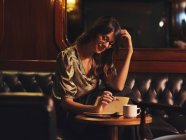 Pensive charmante femme dans des lunettes écrit dans un cahier à la table ronde en bois avec café dans le café — Photo de stock