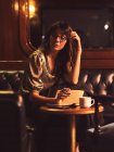 Pensive charming woman in glasses writing in notebook at round wooden table with coffee in cafe — Stock Photo
