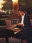 Mujer de negocios inteligente en gafas de escribir en el ordenador portátil y comprobar la información en el teléfono móvil cómodamente sentado en el sofá de cuero negro en la cafetería - foto de stock