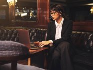 Kluge Geschäftsfrau mit Brille tippt auf Laptop und sitzt gemütlich auf schwarzem Ledersofa im Café — Stockfoto