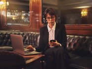 Mulher de negócios inteligente em óculos digitando no laptop e verificando informações no telefone móvel confortavelmente sentado no sofá de couro preto no café — Fotografia de Stock