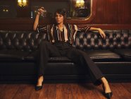 Fashionable stylish woman in trendy clothes holding glass of alcohol sitting wide spread apart legs on black leather couch and looking at camera — Stock Photo