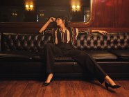 Modisch stilvolle Frau in trendigen Klamotten sitzt breitbeinig auf schwarzem Ledersofa und schaut weg — Stockfoto