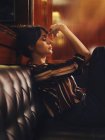 Модная стильная женщина в модной одежде сидит на черном кожаном диване и смотрит в сторону — стоковое фото