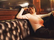 Модна стильна жінка в модному одязі, сидячи на чорному шкіряному дивані і дивлячись — стокове фото