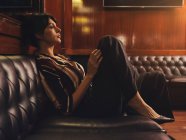 Модна стильна жінка в модному одязі, сидячи на чорному шкіряному дивані і дивлячись — стокове фото