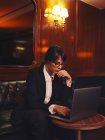 Розумна бізнес-леді в окулярах, друкуючи на ноутбуці, зручно сидить у чорному шкіряному дивані в кафе — стокове фото