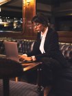 Femme d'affaires intelligente en lunettes tapant sur un ordinateur portable confortablement assis dans un canapé en cuir noir dans un café — Photo de stock