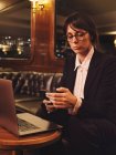 Kluge Geschäftsfrau mit Brille tippt auf Laptop und checkt Informationen im Handy bequem auf schwarzem Ledersofa im Café — Stockfoto