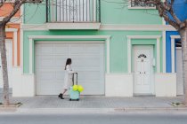 Side view of young stylish female with yellow balloons and suitcase walking on city street next to old styled colorful building — Stock Photo