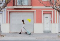 Seitenansicht der jungen stilvollen Frau mit gelben Luftballons und Koffer zu Fuß auf der Straße neben alten bunten Gebäude im Stil — Stockfoto