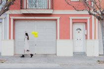 Боковой вид на неузнаваемое стильное женское покрывающее лицо с желтыми воздушными шарами, идущее с чемоданом на городской улице рядом со старым стильным красочным зданием — стоковое фото