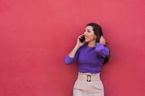 Positivo giovane femmina in camicetta viola e pantaloni beige chiaro parlando sul telefono cellulare mentre in piedi contro colorato sfondo muro rosso — Foto stock