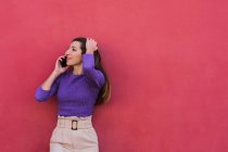 Positive junge Frau in violetter Bluse und hellbeiger Hose, die mit dem Handy telefoniert, während sie vor buntem roten Wandhintergrund steht — Stockfoto