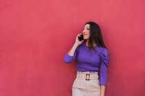 Позитивная молодая женщина в фиолетовой блузке и светло-бежевых брюках разговаривает по мобильному телефону, стоя на фоне красочной красной стены — стоковое фото
