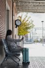 Vista lateral de la joven viajera femenina con estilo con maleta sentada en el banco y esperando el transporte en la estación de tren en el día soleado - foto de stock
