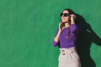 Jovem fêmea positiva em blusa violeta e calças bege claro falando no telefone celular, enquanto em pé contra fundo de parede verde colorido — Fotografia de Stock