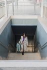 From above of elegant stylish young female traveler with suitcase walking up stairs at train station — Stock Photo