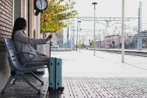 Vista lateral de la joven viajera femenina con estilo con maleta sentada en el banco y esperando el transporte en la estación de tren en el día soleado - foto de stock