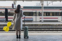 Обратный вид на неузнаваемую женщину-путешественницу в стильном наряде, стоящую с желтыми воздушными шарами в руке и чемоданом на платформе железнодорожного вокзала — стоковое фото