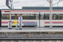 Joven viajera con elegante atuendo de pie con globos amarillos en la mano y maleta en la plataforma de la estación de tren - foto de stock