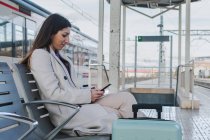 Vue latérale du voyageur féminin moderne avec une valise assise sur un banc sur le quai de la gare et utilisant un smartphone en attendant le train — Photo de stock
