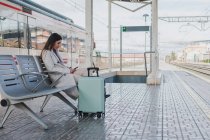 Vista lateral do viajante moderno feminino com mala sentada no banco na plataforma da estação ferroviária e usando o smartphone enquanto espera pelo trem — Fotografia de Stock