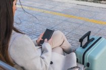 Сверху лопнула неузнаваемая путешественница с чемоданом, сидящим на скамейке на платформе железнодорожного вокзала и пользующимся смартфоном в ожидании поезда — стоковое фото
