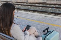De arriba copped viajero femenino irreconocible con maleta sentado en el banco en la plataforma de la estación de tren y el uso de teléfono inteligente mientras espera el tren - foto de stock