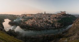 Panoramic view across river of old city Toledo in Spain with medieval castles and fortresses at sunset time with cloudy sky and reflection in river water — Stock Photo