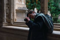 Vista laterale del viaggiatore maschio con zaino in piedi accanto alla finestra e scattare foto con la fotocamera durante la visita del vecchio edificio storico in pietra nella città spagnola Toledo — Foto stock