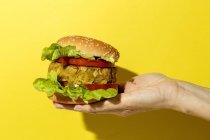 Cortada mano irreconocible persona sosteniendo una hamburguesa de lenteja verde vegana casera con tomate, lechuga y papas fritas sobre un fondo colorido amarillo - foto de stock