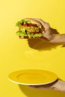 Cropped unrecognizable person hand holding a homemade vegan green lentil burger with tomato, lettuce and french fries on a yellow colorful background — Stock Photo