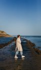 Femme en vêtements décontractés et grand chien mastiff brun regardant la caméra souriant tout en marchant le long de jetée en bois humide contre l'eau calme de la baie sous le ciel bleu en Espagne — Photo de stock