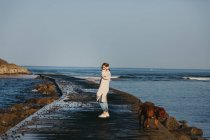 Vista lateral de la mujer viajera en ropa casual caminando por el muelle de madera húmeda contra las olas del mar y la costa rocosa en España - foto de stock