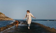 Female in casual clothes and big brown Mastiff dog looking at each other while walking along wet wooden pier against calm bay water under blue sky in Spain — Stock Photo