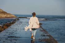 Vista trasera de una viajera sin rostro vestida con ropa casual caminando por el muelle de madera mojada contra las olas del mar y la costa rocosa en España - foto de stock