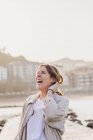Mujer rubia feliz en ropa casual con los ojos cerrados riendo mientras está de pie en el muelle y tocando el pelo contra el ambiente urbano borroso de la ciudad turística en España - foto de stock
