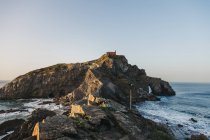 Paving stone way leading along stone bridge and ridge of rocky hill to lonely country house on island Gaztelugatxe surrounded by tranquil sea water with white foam waves — Stock Photo