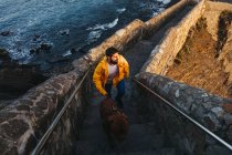 High angle of male in bright yellow jacket with big brown dog walking up stone stairs and looking away with interest against troubled bay water washing rocky coast in Spain during sunrise — Stock Photo