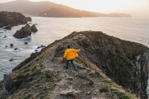 Обратный вид на неузнаваемого человека в яркой желтой куртке и джинсе, бегущего по скалистому холму и наслаждающегося живописными пейзажами морского побережья во время заката в Испании — стоковое фото