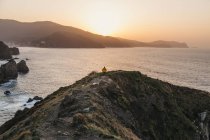 Vue arrière d'un homme méconnaissable en veste jaune vif et en denim courant sur une colline rocheuse et profitant d'un paysage pittoresque de la côte de la mer pendant le coucher du soleil en Espagne — Photo de stock