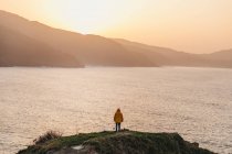 Обратный вид на неузнаваемого человека в яркой желтой куртке и джинсе, стоящего на скалистом холме и наслаждающегося живописными пейзажами морского побережья во время заката в Испании — стоковое фото