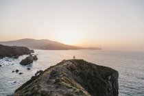 Vista trasera de un hombre irreconocible con chaqueta amarilla vibrante y denim parado en una colina rocosa y disfrutando de pintorescos paisajes de la costa del mar durante la puesta de sol en España - foto de stock