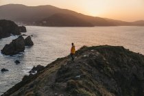 Vista lateral do homem irreconhecível com jaqueta amarela vibrante e ganga em pé na colina rochosa e desfrutando de paisagens pitorescas da costa do mar durante o pôr do sol na Espanha — Fotografia de Stock