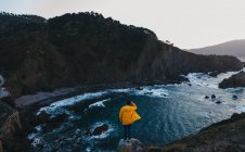 Von oben Rückenansicht einer Person in leuchtend gelber Jacke, die am Rande der Klippe steht und die atemberaubende Landschaft der felsigen Meeresküste während des Sonnenuntergangs in Spanien genießt — Stockfoto