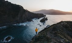 Desde arriba vista trasera de la persona en chaqueta amarilla vibrante de pie en el borde del acantilado y disfrutar de un paisaje increíble de la costa rocosa del mar durante la puesta de sol en España - foto de stock