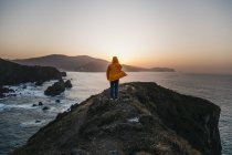 Visão traseira do homem irreconhecível com jaqueta amarela vibrante e ganga em pé na colina rochosa e desfrutando de paisagem pitoresca da costa do mar durante o pôr do sol na Espanha — Fotografia de Stock