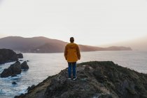 Vista trasera de un hombre irreconocible con chaqueta amarilla vibrante y denim parado en una colina rocosa y disfrutando de pintorescos paisajes de la costa del mar durante la puesta de sol en España - foto de stock